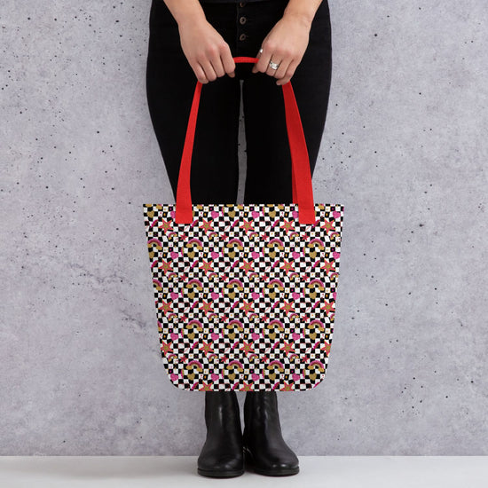 A person in black pants and boots holds a Galaxy Tote Bag from My Favourite Colour is Rainbow, featuring a colourful geometric pattern and bright red handles, against a grey speckled background.