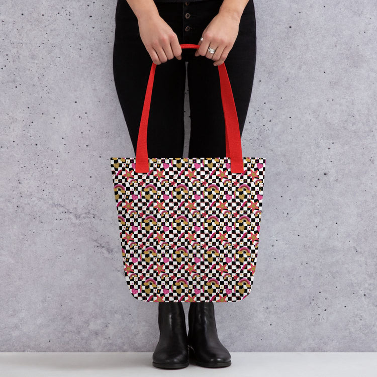 A person in black pants and boots holds a Galaxy Tote Bag from My Favourite Colour is Rainbow, featuring a colourful geometric pattern and bright red handles, against a grey speckled background.