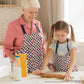 An older woman and a young girl, both wearing My Favourite Colour is Rainbows Galaxy Apron, are baking together. The girl rolls dough on the table while the woman smiles. Milk, eggs, and a mixing bowl rest nearby in the naturally lit kitchen.