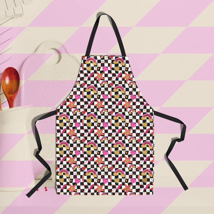 The Galaxy Apron from My Favourite Colour is Rainbow features lively black and white checkered designs with glittery gold, pink and red stars, rainbows and hearts.  The background features cooking utensils.