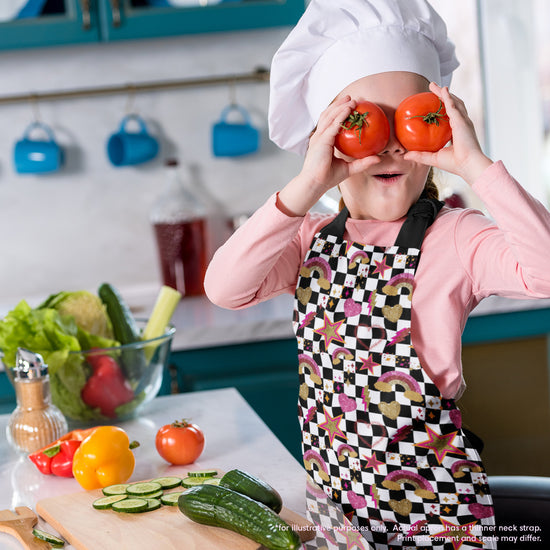 A child in a Galaxy Apron from My Favourite Colour is Rainbow playfully holds two tomatoes over their eyes in a lively kitchen with sliced cucumbers, bell pepper, and lettuce on the counter, while utensils and containers adorn the background.