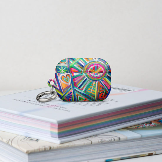 The Geometric Rainbow Airpods Pro Gen 2  Case stands on a stack of books.  It features  an abstract design of colourful green, pink, yellow, blue, and purple stripes, dots, and zigzags. 
