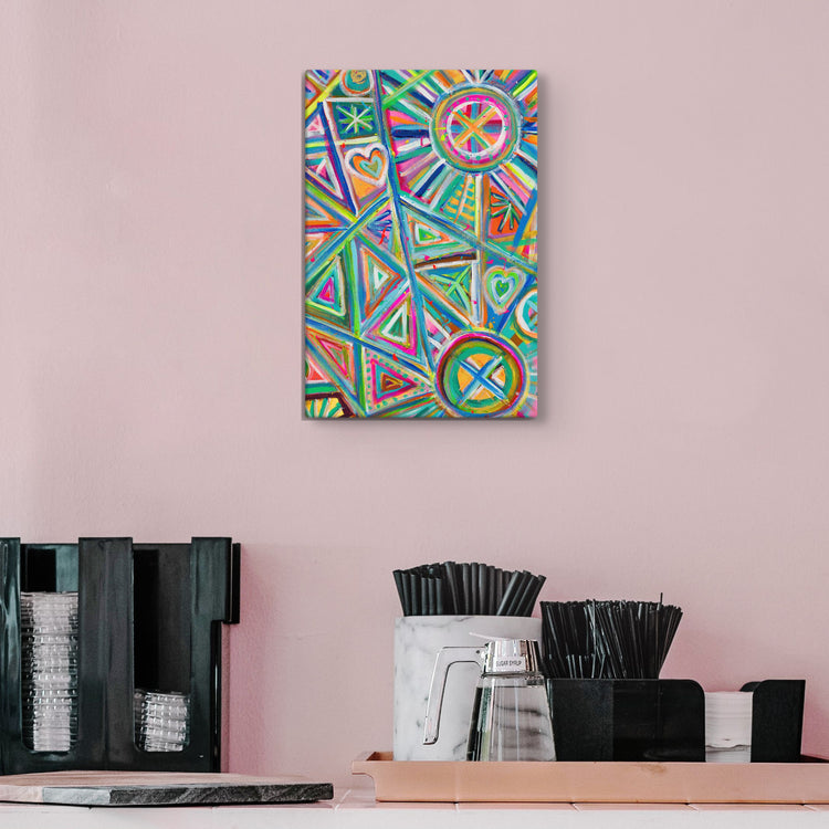 The Geometric Rainbow Canvas Artwork by My Favourite Colour is Rainbow, showcasing vibrant geometric shapes and patterns in various colours, hangs on a pale pink wall. Below it sits a black organiser with glasses and straws next to a clear pitcher.