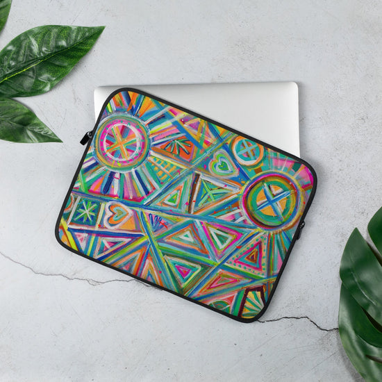 A laptop slips halfway out of the 13" Geometric Rainbow Laptop Sleeve by My Favourite Colour is Rainbow, showcasing colourful patterns like triangles, circles, and hearts. It rests on a light gray surface with nearby green leaves.