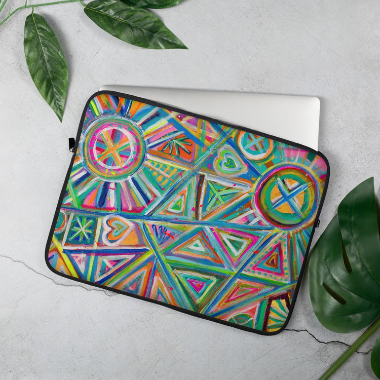 A laptop slips halfway out of the 15" Geometric Rainbow Laptop Sleeve by My Favourite Colour is Rainbow, showcasing colourful patterns like triangles, circles, and hearts. It rests on a light gray surface with nearby green leaves.