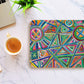 The Geometric Rainbow Mouse Pad by My Favourite Colour is Rainbow, featuring abstract triangles, circles, and hearts, rests on a white marble surface. Nearby are a cup of coffee, a laptop, and a small green plant adding a touch of nature.