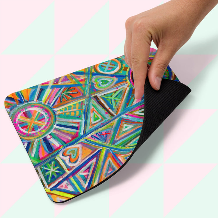 A hand lifts the corner of a colorful Geometric Rainbow Mouse Pad by My Favourite Colour is Rainbow, featuring geometric shapes, hearts, and circular motifs in vibrant blue, orange, and pink against a pale green and pink geometric background.