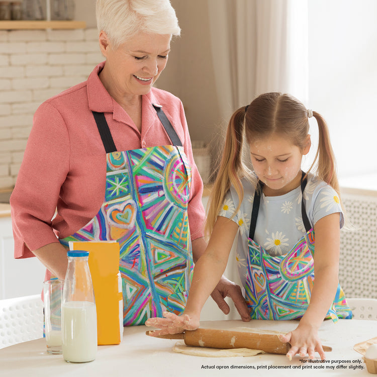 In a bright kitchen, a grandmother and granddaughter bake together while wearing My Favourite Colour is Rainbows durable Geometric Rainbow Aprons. The girl uses a rolling pin as her grandmother smiles warmly. Milk and flour rest on the table nearby.  The aprons feature vivid geometric designs in blue, pink, and green 