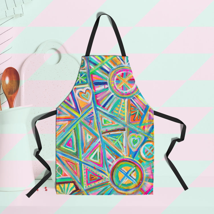 The Geometric Rainbow Apron by My Favourite Colour is Rainbow features a vibrant abstract design with triangles, circles, and hearts in bold colours. The background features cooking utensils and a cutting board.