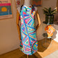 A mannequin displays the Geometric Rainbow Apron by My Favourite Colour is Rainbow, featuring vibrant blue, red, pink, and green circles and triangles. Made from durable high-quality fabric, it complements a plant, a colourful flower, and modern art on the walls.