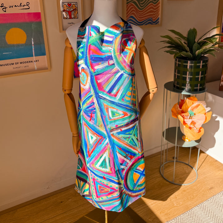 A mannequin displays the Geometric Rainbow Apron by My Favourite Colour is Rainbow, featuring vibrant blue, red, pink, and green circles and triangles. Made from durable high-quality fabric, it complements a plant, a colourful flower, and modern art on the walls.