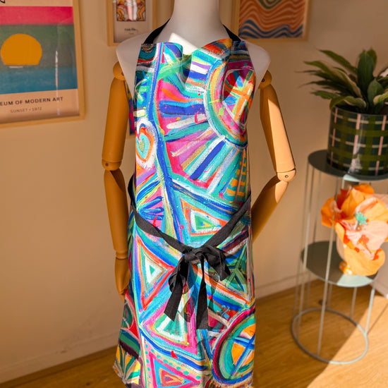 A mannequin displays the Geometric Rainbow Apron by My Favourite Colour is Rainbow in a well-lit room. The durable apron features vivid geometric designs in blue, pink, and green, surrounded by a table with a potted plant and colourful framed artwork on the wall.