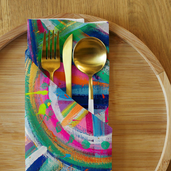 The Geometric Rainbow Cloth Napkin Set of 4 by My Favourite Colour is Rainbow holds gold utensils—a fork, knife, and spoon—on a wooden plate. The napkin features vibrant swirls and splatters of pink, blue, green, and yellow for an eye-catching dining upgrade.