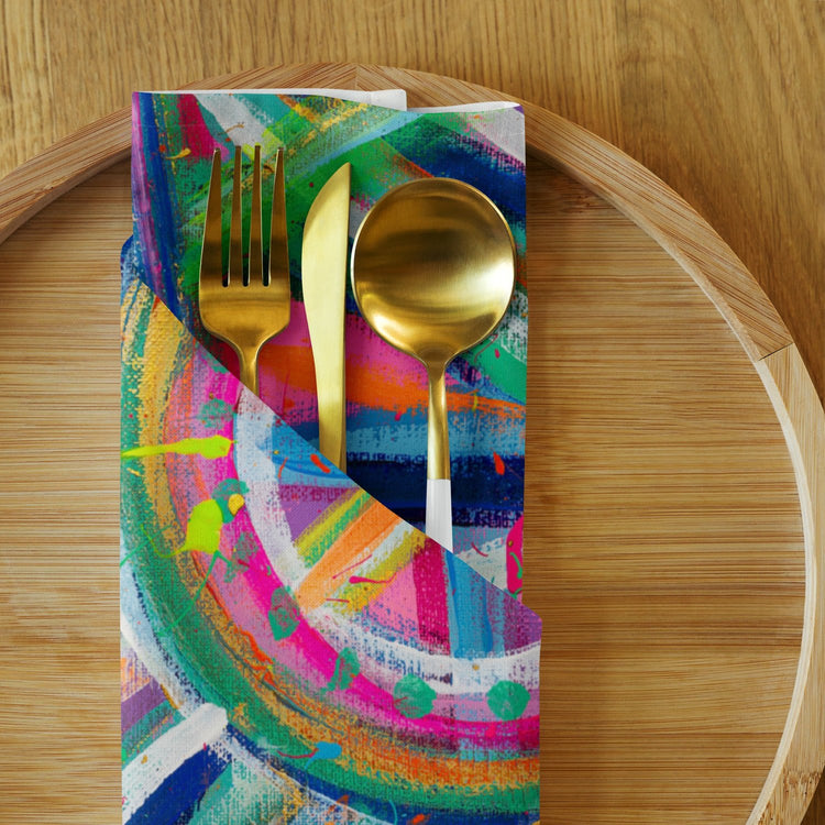 The Geometric Rainbow Cloth Napkin Set of 4 by My Favourite Colour is Rainbow holds gold utensils—a fork, knife, and spoon—on a wooden plate. The napkin features vibrant swirls and splatters of pink, blue, green, and yellow for an eye-catching dining upgrade.