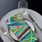 Enhance your modern table with a dining set featuring a white plate with brown edges, a Geometric Rainbow Cloth Napkin by My Favourite Colour is Rainbow, silver cutlery on a textured placemat, an empty wine glass—all stylishly arranged on a dark tabletop.