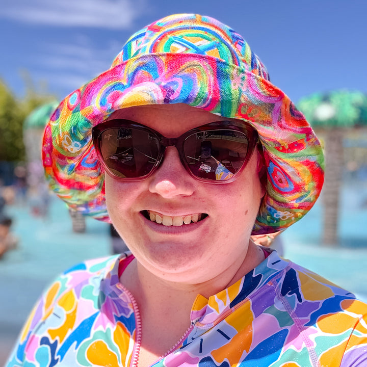 A person wearing a colorful floral shirt, Geometric Rainbow/Love in Neon Reversible Bucket Hat by My Favourite Colour is Rainbow, and sunglasses smiles at the camera with a water area, people, and trees under a blue sky in the background.