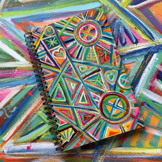 The Geometric Rainbow Notebook from My Favourite Colour is Rainbow, featuring a colourful spiral-bound cover with geometric patterns of triangles, circles, and hearts in vibrant blue, green, pink, and orange hues, rests on a vibrant abstract background of the same painting.