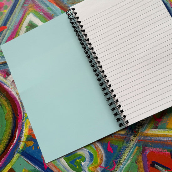 The My Store Geometric Rainbow Notebook showcases an open spiral design with a blank blue cover and lined pages. Set against an abstract backdrop, it radiates creativity through vivid shades and geometric patterns.