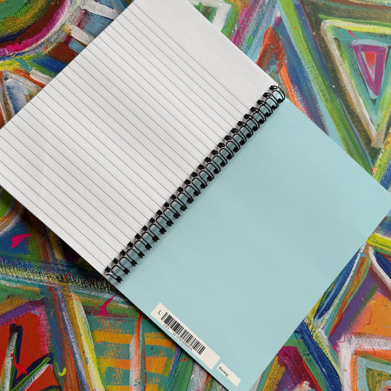 A Geometric Rainbow Notebook by My Store, featuring lined pages and a light blue cover, rests on a vibrant abstract surface. Its colorful patterns inspire creativity as the barcode peeks from the corner.