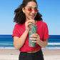 A woman with long curly hair, wearing red sunglasses and a pink shirt, smiles while sipping from a Geometric Rainbow Stainless Steel Tumbler by My Favourite Colour is Rainbow, using a metal straw. She stands on a beach with blue ocean waves and clear skies in the background.