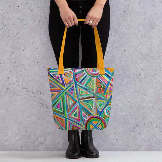 A person holds the Geometric Rainbow Tote Bag by My Favourite Colour is Rainbow. The spacious tote features colourful geometric and heart patterns in vibrant green, blue, orange, and pink. These bright hues pop against their black pants and ankle boots.