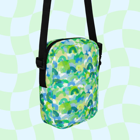 The Green Rainbow Mini Crossbody Bag by My Favourite Colour is Rainbow showcases a vibrant print with abstract blue, green, and white waves over a pale green and white checkerboard. It includes a black adjustable strap for hands-free use.