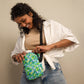 A curly-haired person in a white shirt and jeans smiles while adjusting a Green Rainbow Mini Crossbody Bag by My Favourite Colour is Rainbow, enjoying its hands-free convenience against a plain, neutral background.