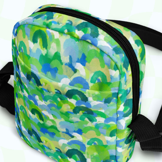 The Green Rainbow Mini Crossbody Bag by My Favourite Colour is Rainbow showcases a vibrant print in green, blue, and white with a pale green checkered background. It features a front pocket, black zippers, and padded black shoulder straps for hands-free convenience.