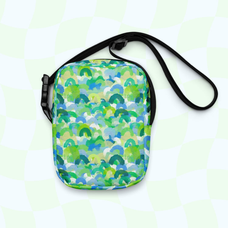 The Green Rainbow Mini Crossbody Bag from My Favourite Colour is Rainbow showcases green, blue, and white shades on a checkered light green backdrop. The bag offers hands-free convenience with its black strap.