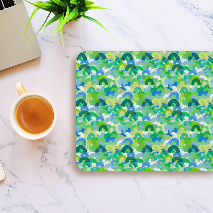 A coffee cup rests on a marble surface beside a My Favourite Colour is Rainbows Green Rainbow Mouse Pad, featuring a green, blue, and white abstract pattern. A corner of a laptop and a small potted plant are partially visible at the top left.