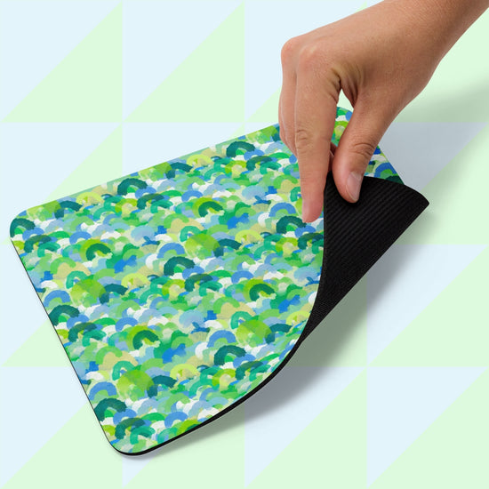 A hand lifts the corner of the Green Rainbow Mouse Pad by My Favourite Colour is Rainbow, featuring a colorful abstract pattern in green and blue. The back is black, revealing a light green geometric background.