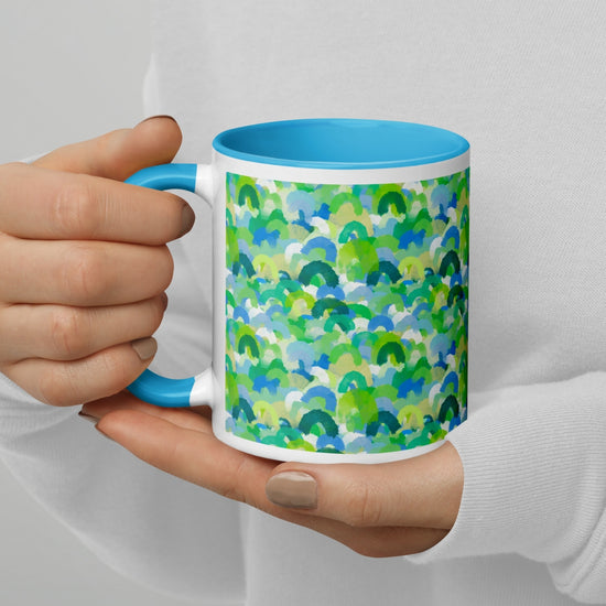 A person with light skin, wearing a white sweater and light brown nail polish, holds the Green Rainbow Mug by My Favourite Colour is Rainbow. The ceramic mug features a blue interior and handle with an abstract pattern of green, blue, and turquoise watercolor-like shapes.