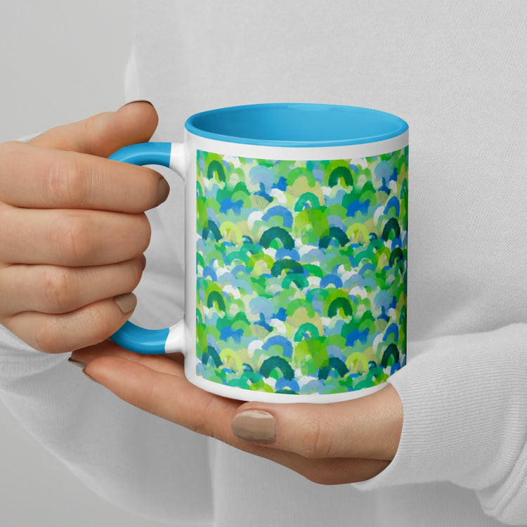 A person with light skin, wearing a white sweater and light brown nail polish, holds the Green Rainbow Mug by My Favourite Colour is Rainbow. The ceramic mug features a blue interior and handle with an abstract pattern of green, blue, and turquoise watercolor-like shapes.