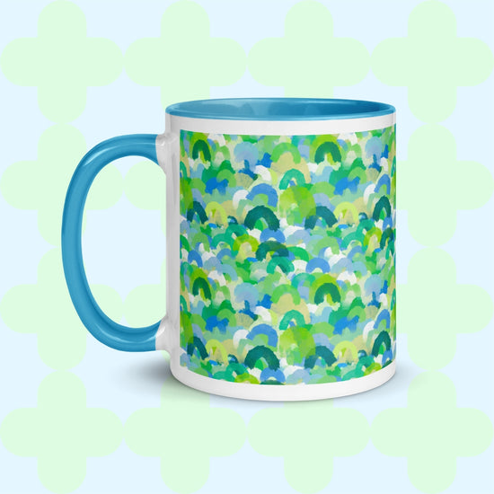 The Green Rainbow Mug by My Favourite Colour is Rainbow is a ceramic mug featuring a blue handle and rim, decorated with vibrant green, blue, and teal brushstroke designs. Its light background showcases watercolor rainbows of abstract soft green shapes for an artistic touch.