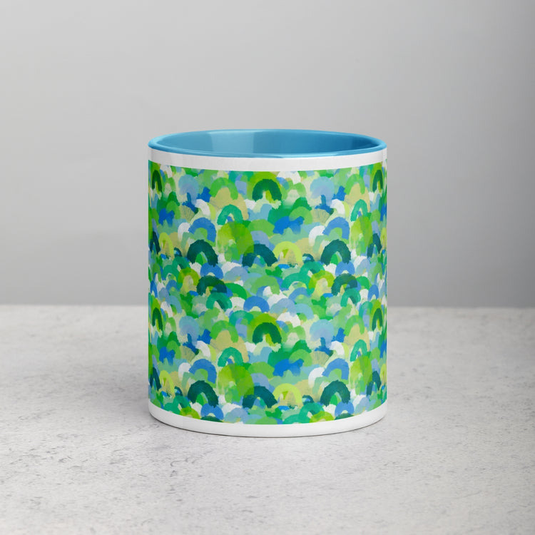 The Green Rainbow Mug by My Favourite Colour is Rainbow showcases an abstract pattern of overlapping green, blue, and turquoise shapes evoking watercolor rainbows or ocean waves. Its ceramic with a vibrant blue interior, elegantly displayed on a light grey surface against a matching background.