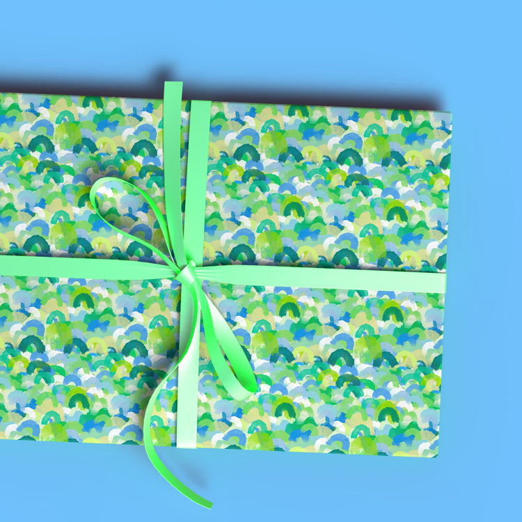 A rectangular gift box wrapped in My Favourite Colour is Rainbows Green Rainbow Wrapping Paper Sheets features blue, green, and white abstract designs with a green ribbon on a light blue background.