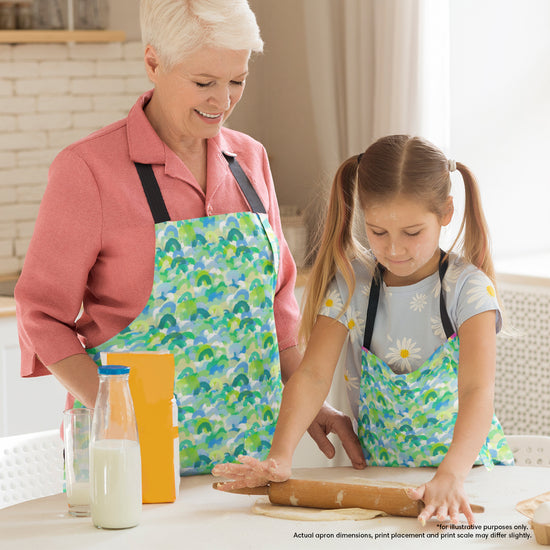 A woman and young girl, both in matching Green Rainbow Aprons by My Favourite Colour is Rainbow, are baking together. The girl rolls dough with a rolling pin on a table surrounded by ingredients like milk and flour.  The aprons feature overlapping rainbows in shades of green and blue.