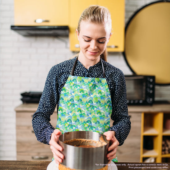 A woman in a Green Rainbow Apron by My Favourite Colour is Rainbow stands in a kitchen, holding a springform pan with crust. She is focused on her task amid yellow cabinets and appliances, creating a cosy cooking atmosphere. The apron features overlapping rainbows in shades of green and blue.