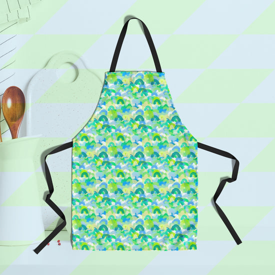 A My Favourite Colour is Rainbows Green Rainbow Apron, featuring overlapping rainbows in shades of green, blue, and white, is displayed on a light green and white checkered background with a partially visible white cutting board and spoons.