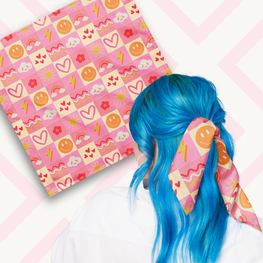 Two views of the 'Happy Smiles Sunny Days' Bandana by My Favourite Colour is Rainbow.  At top left, a flat lay of the bandana.  At bottom right, a person with vibrant blue hair is seen from behind, wearing the bandana in her hair.  The bandana features a vibrant pattern of smiley faces, lightning bolts, hearts, clouds, flowers, and waves on a pink and cream checkered background.