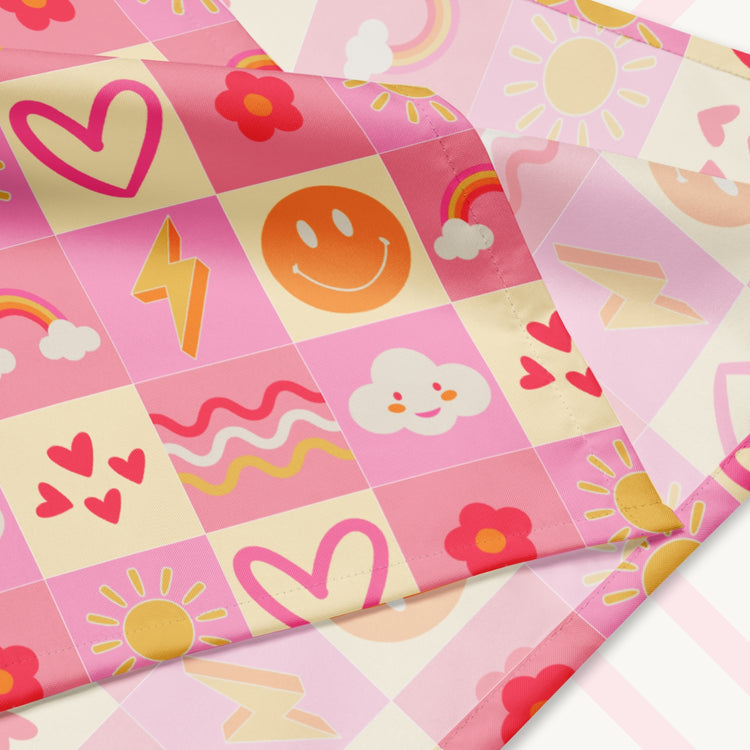 A closeup of the 'Happy Smiles Sunny Days' Bandana by My Favourite Colour is Rainbow featuring a vibrant pattern of smiley faces, lightning bolts, hearts, clouds, flowers, and waves on a pink and cream checkered background. The design is printed on only one side of the bandana.