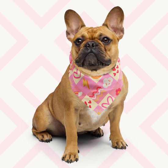 A brown French Bulldog with an attentive expression sits against a light geometric background, wearing the lightweight Happy Smiles Sunny Days Bandana by My Favourite Colour is Rainbow. The bandana features a vibrant pattern of smiley faces, lightning bolts, hearts, clouds, flowers, and waves on a pink and cream checkered background.