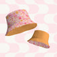 Two Happy Smiles Sunny Days reversible bucket hats by My Favourite Colour is Rainbow float in mid-air. One hat has emojis, hearts, and peace signs on pink; the other is solid orange. Both wide-brimmed hats are set against a pink and white geometric background.