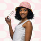 A curly-haired woman, wearing the Happy Smiles Sunny Days Reversible Bucket Hat by My Favourite Colour is Rainbow and a white sleeveless top, smiles as she holds glasses. The background displays a pink and white geometric design, ideal for sunny days.