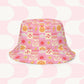 The Happy Smiles Sunny Days Reversible Bucket Hat by My Favourite Colour is Rainbow showcases a pink pattern with smiley faces, hearts, lightning bolts, flowers, sun, and clouds in orange, yellow, and white squares. It features a matching abstract design with similar curved shapes. Perfect for sunny days!.