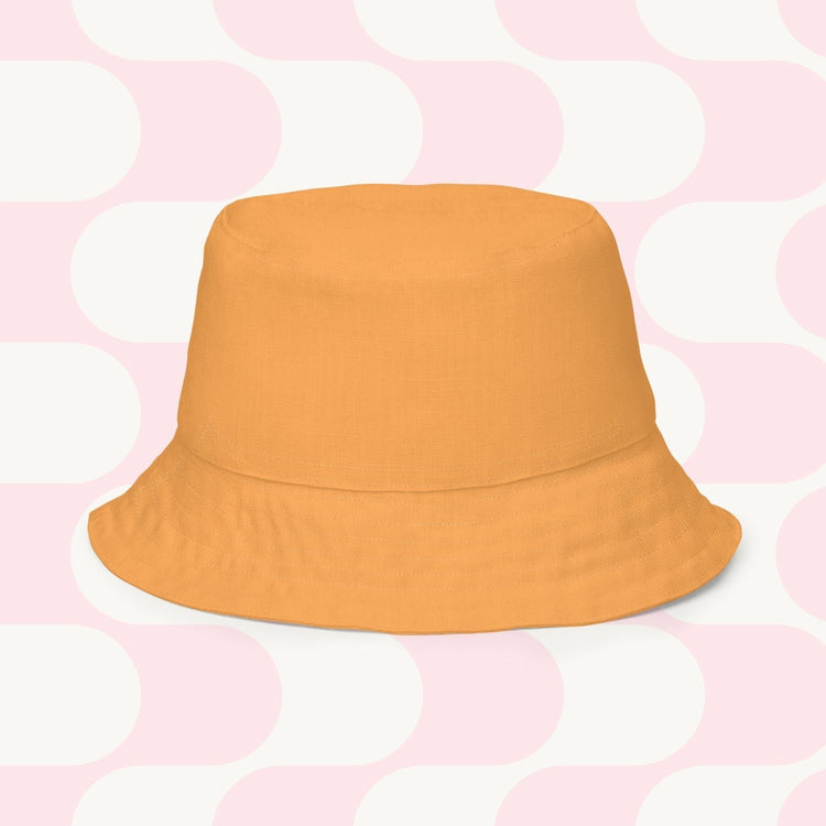 The Happy Smiles Sunny Days Reversible Bucket Hat by My Favourite Colour is Rainbow is set against alternating light pink and white geometric shapes, evoking sunny days and happy smiles with its vibrant orange hue.