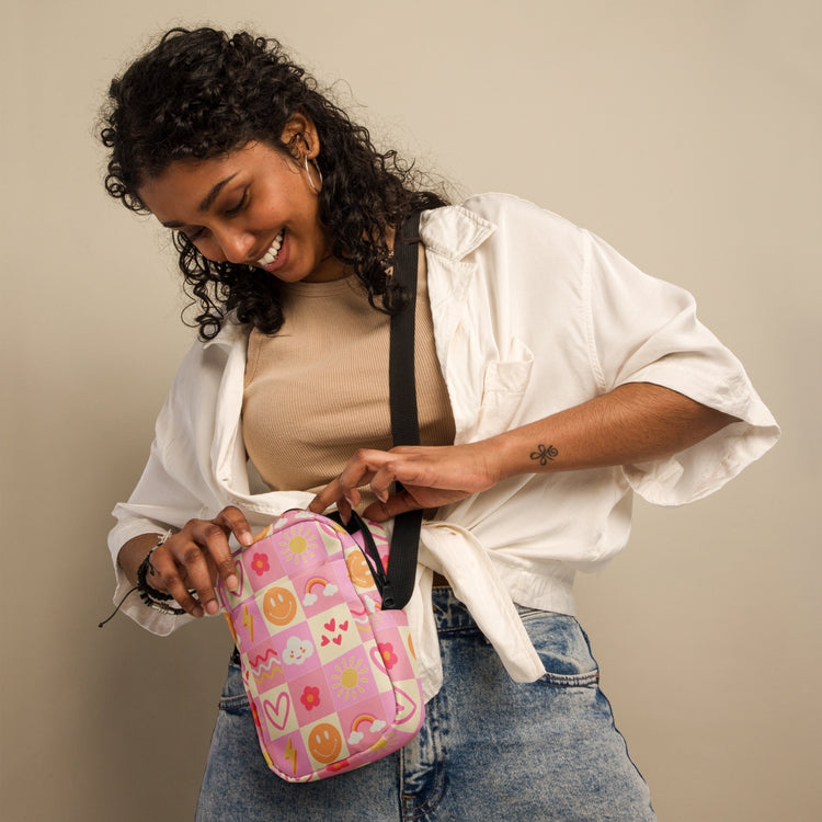 A smiling woman with curly hair wears a beige top, white shirt, and jeans as she looks inside a Happy Smiles Sunny Days Mini Crossbody Bag by My Favourite Colour is Rainbow. The pink checkered bag has playful designs including hearts, smiley faces, rainbows, suns, flowers, and clouds.  It has internal and external pockets and a black strap with a clip on one side that can be extended to crossbody length.