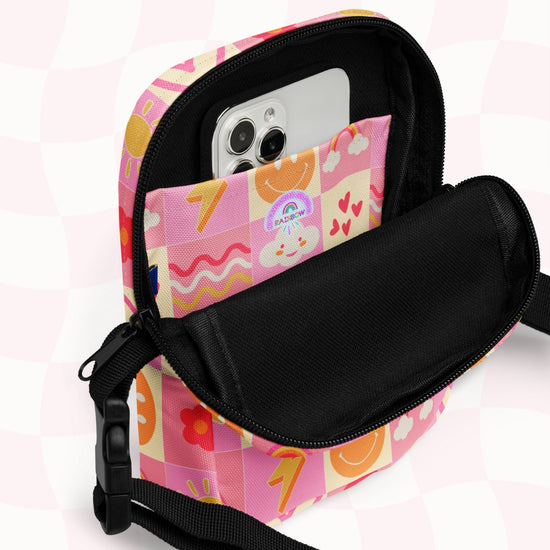 The Happy Smiles Sunny Days Mini Crossbody Bag by My Favourite Colour is Rainbow features a playful multicoloured pattern with hearts, numbers, and a smiling cloud. It includes adjustable straps and an open pocket revealing a smartphone inside.