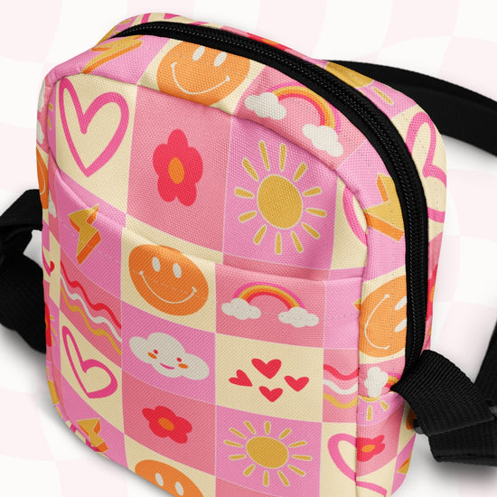 The Happy Smiles Sunny Days Mini Crossbody Bag by My Favourite Colour is Rainbow is a small, pink checkered bag with playful designs including hearts, smiley faces, rainbows, suns, flowers, and clouds. It features black zippers and adjustable straps for a sturdy yet vibrant look.