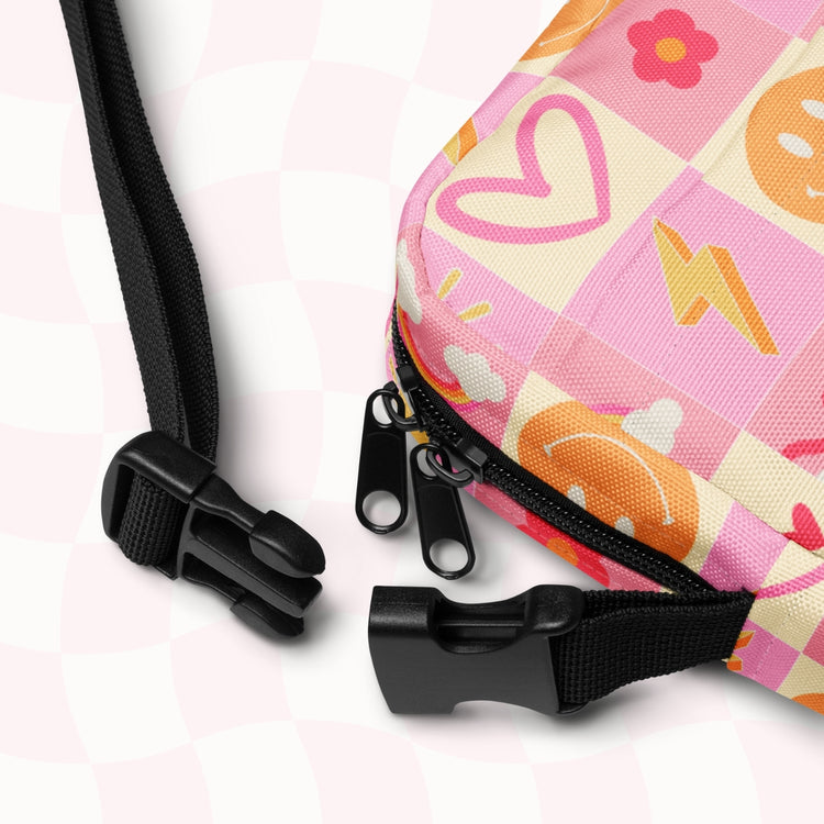 A close-up of the Happy Smiles Sunny Days Mini Crossbody Bag by My Favourite Colour is Rainbow, showcasing a pink and orange design with hearts, lightning bolts, and smiley faces. It features a sturdy zipper with dual pulls, black adjustable straps with a plastic buckle against a light pink checkered background.